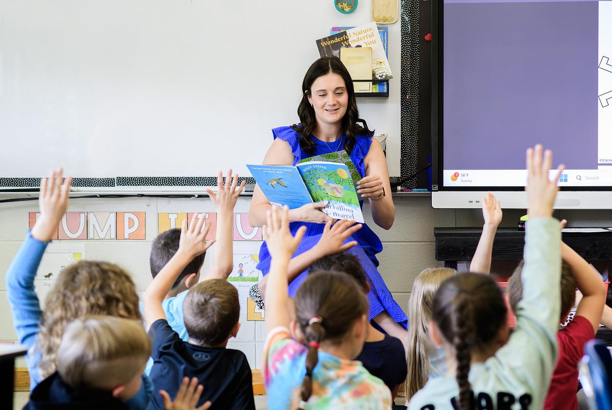 Teacher reads to kids in class with children raising their hands