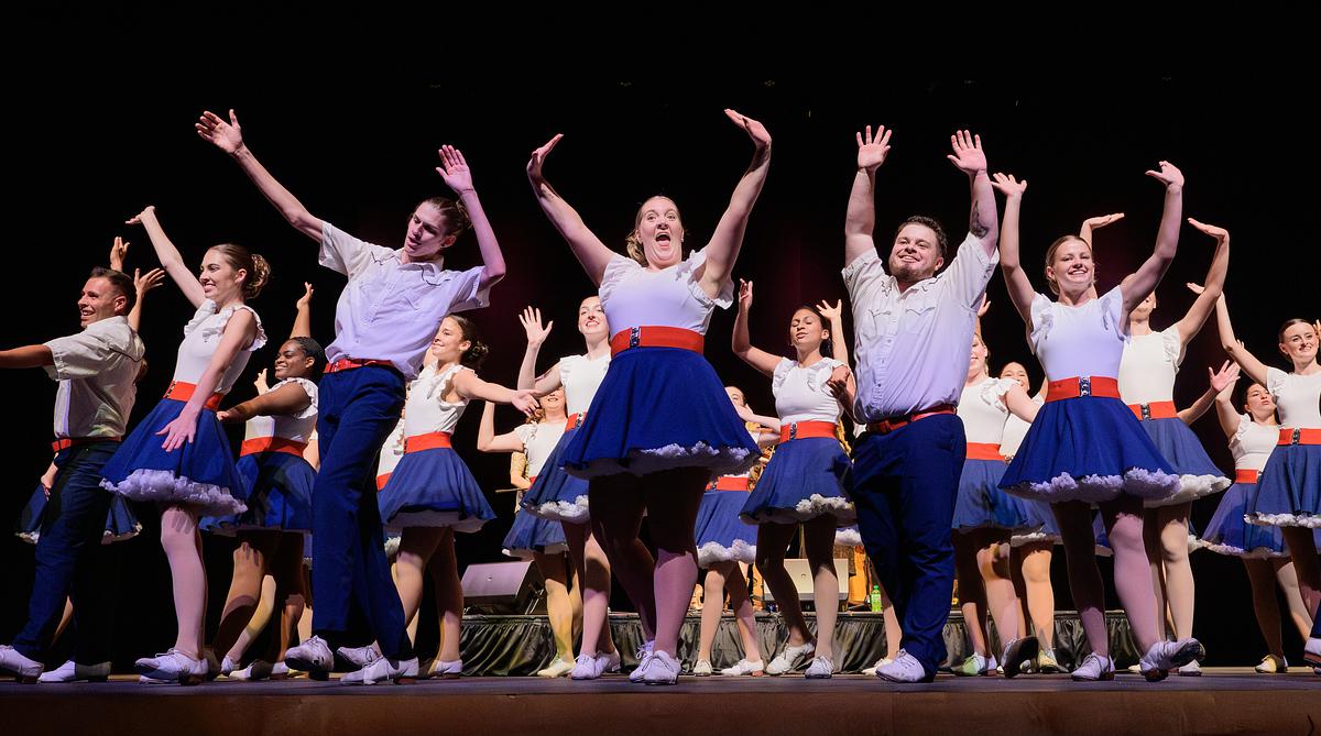 Group of dancers in navy skirts and light purple tops performing enthusiastically on stage with arms raised.