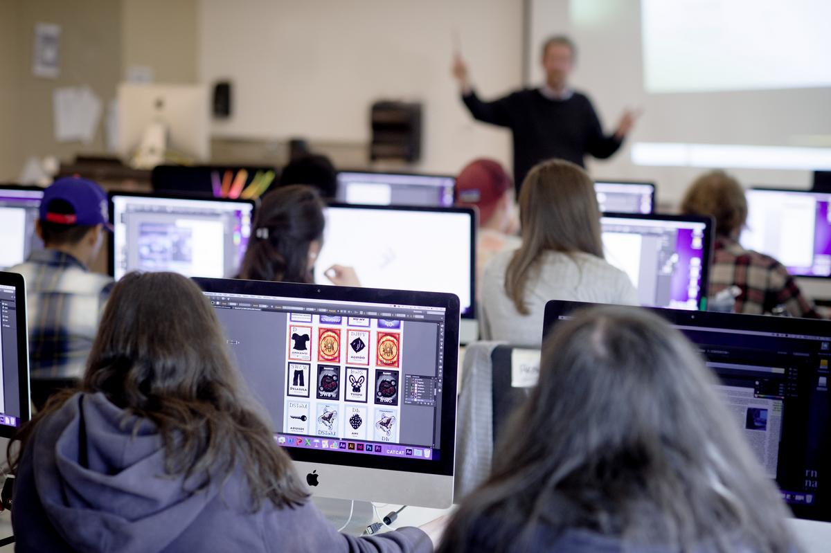 Students sitting at desks on Mac Desktop computers listening to a professor teach graphic design concepts