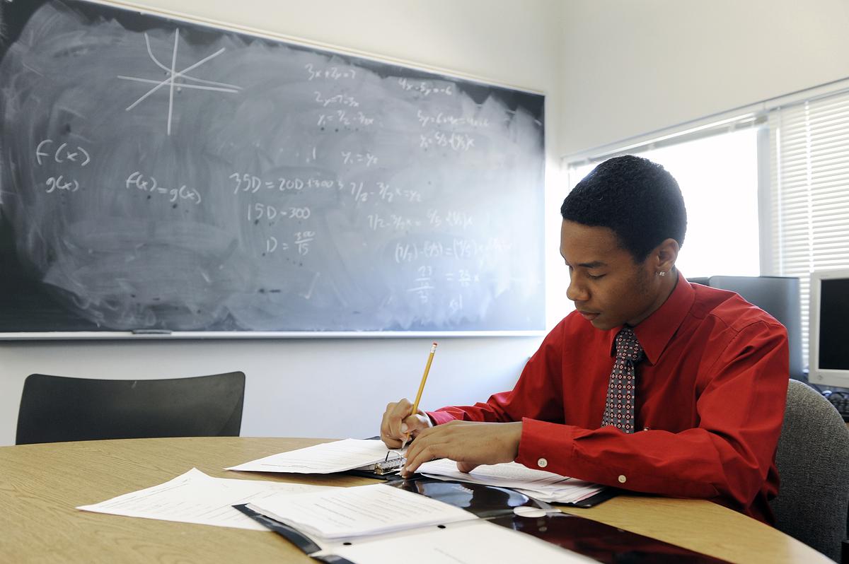 Student in a button-up shirt and tie writes at a desk with math equations written on a chalkboard behind them