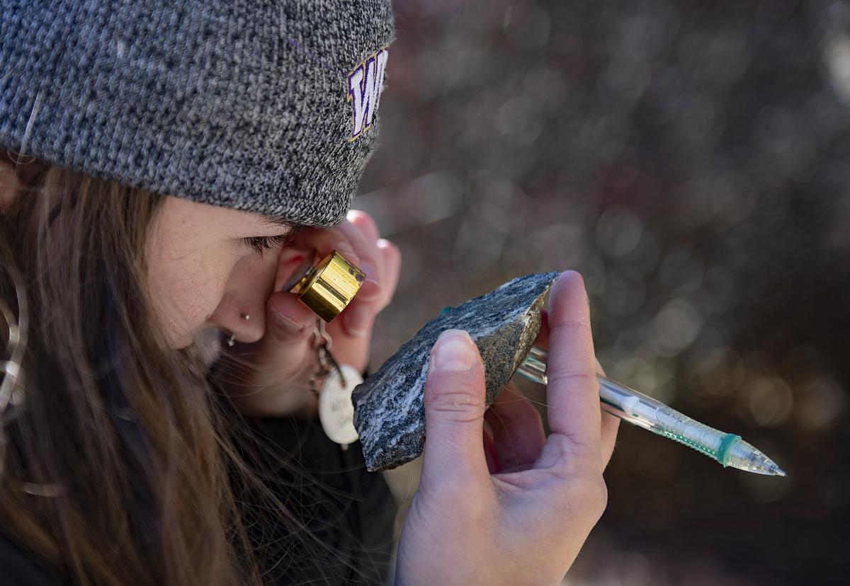 A student standing outside holding a magnifying lens as they study a rock and its composition 
