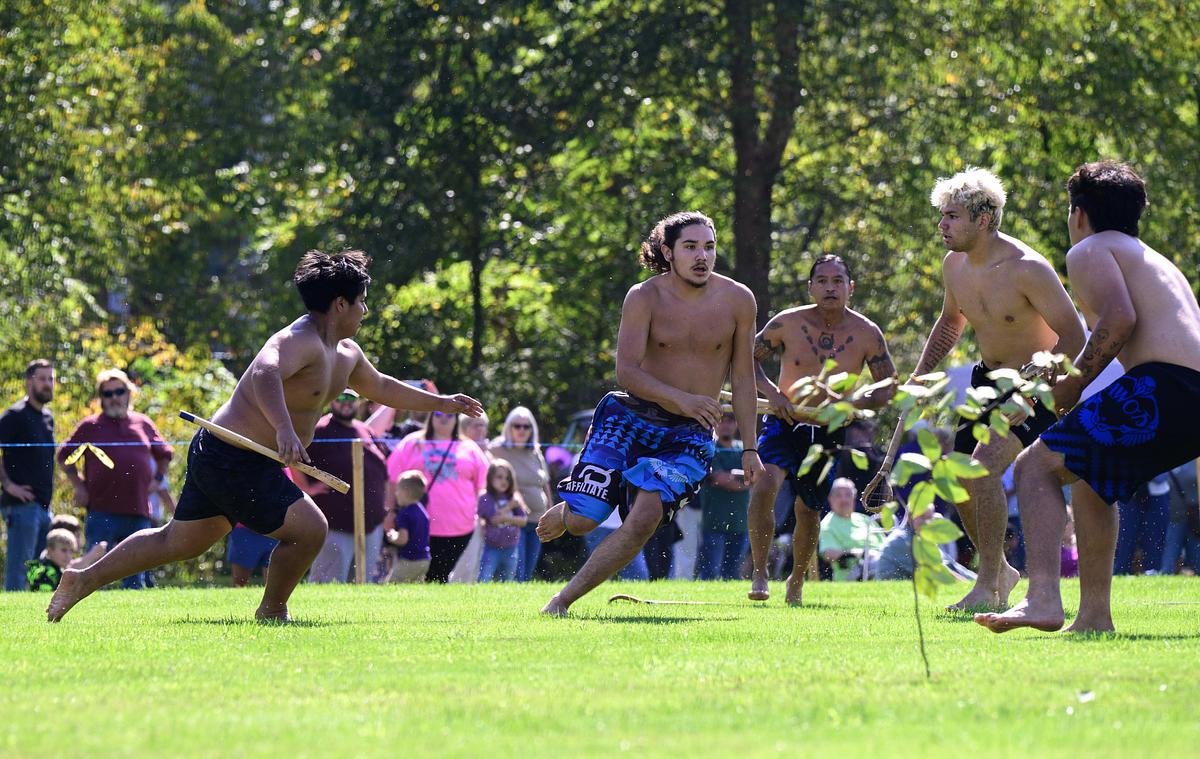 Shirtless men playing stickball on green grass with spectators watching in background