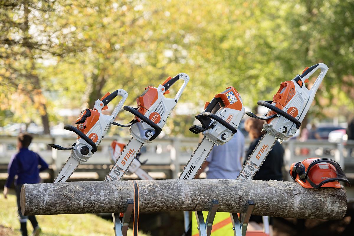Four orange and white chainsaws mounted on a horizontal log with blurred park background and people.