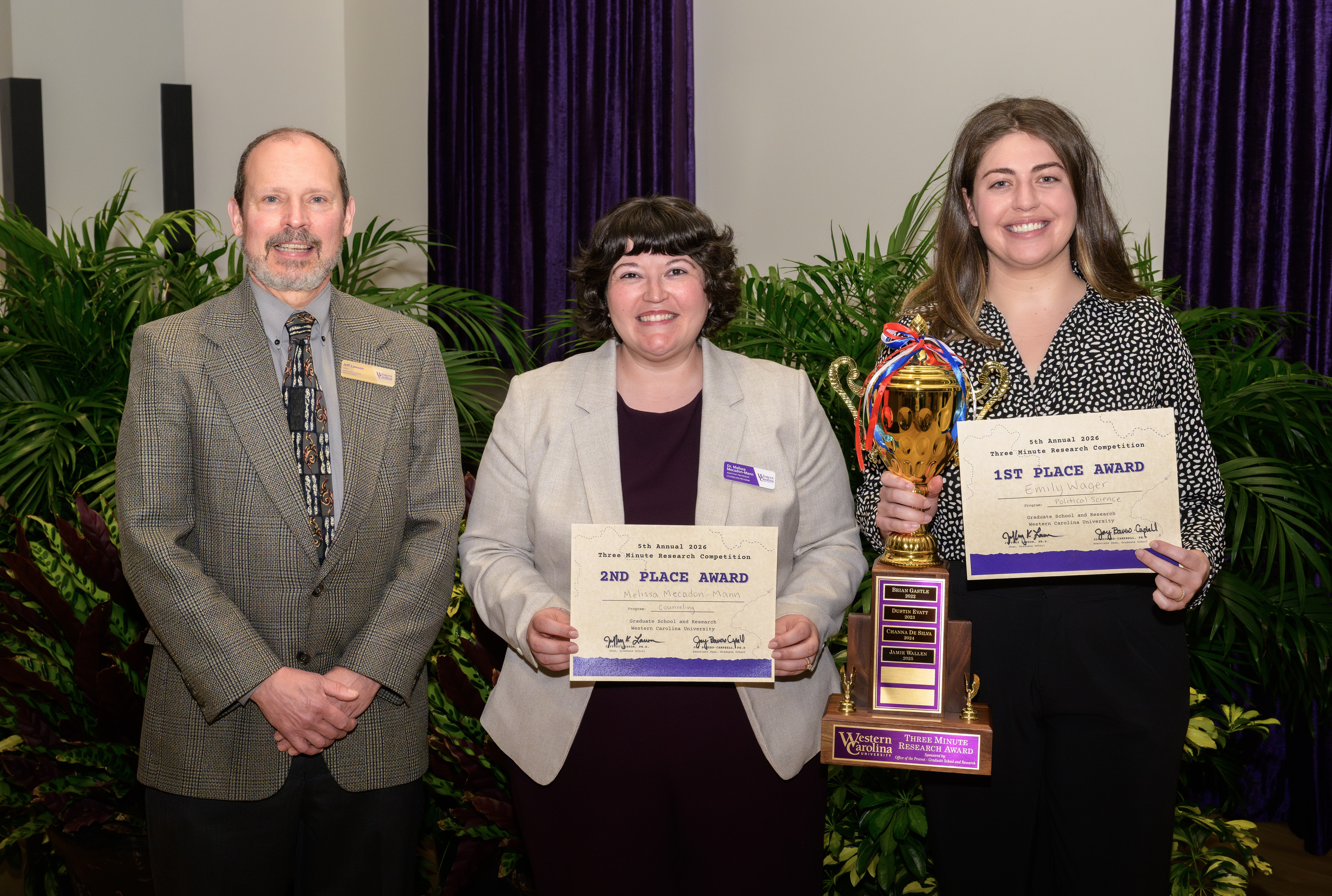 Three people pose with award certificates and a trophy at a formal recognition ceremony.