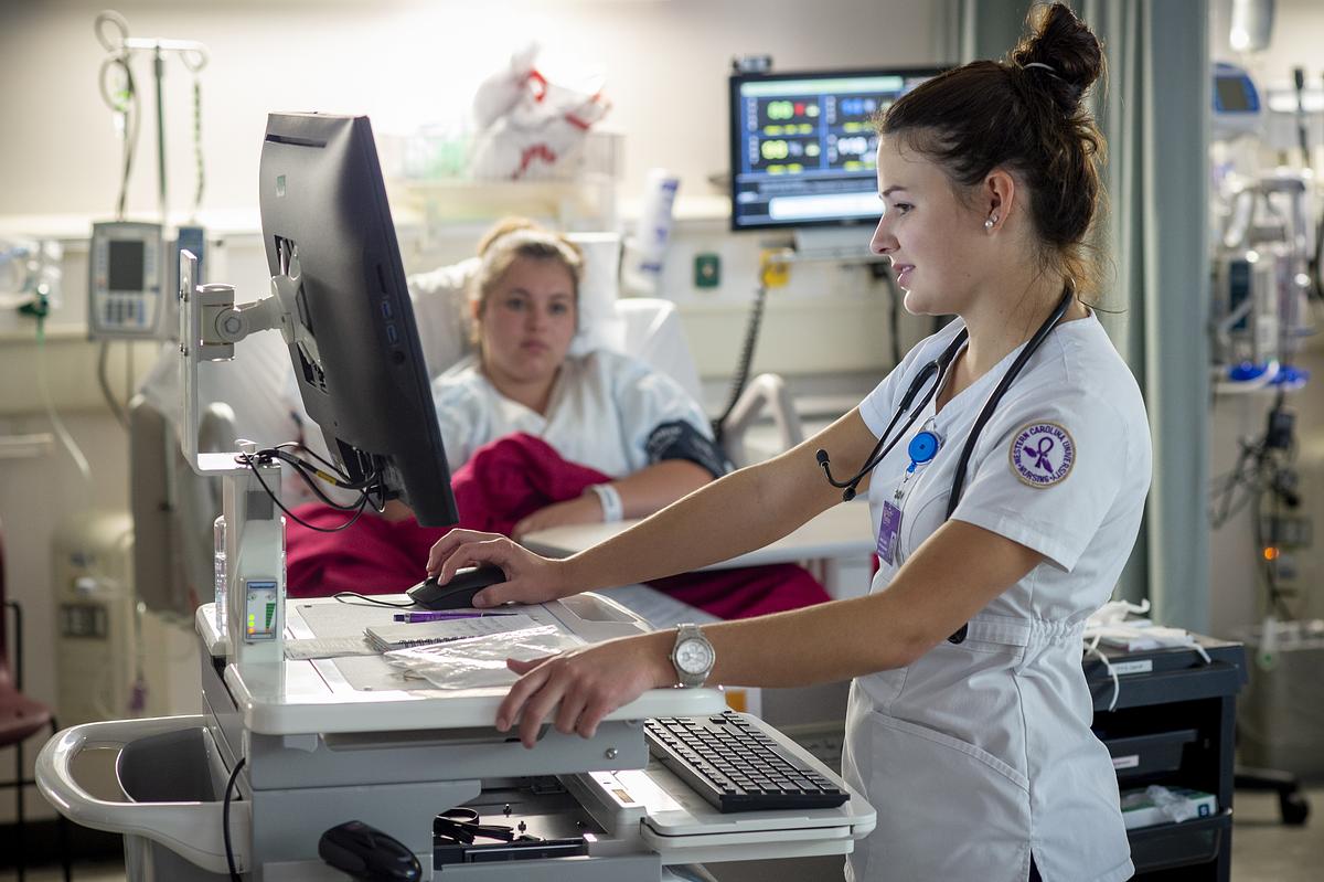 Healthcare professional in white scrubs operates medical monitoring equipment in hospital room with colleague present.