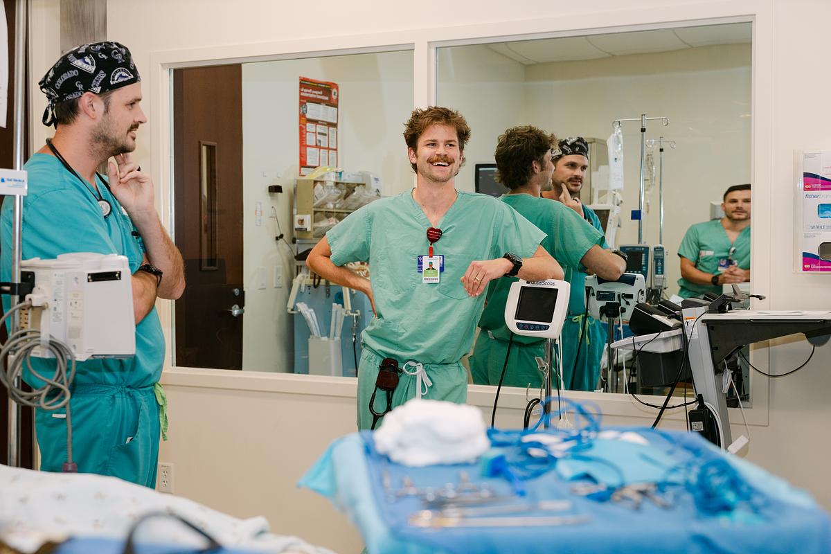 Healthcare workers in medical scrubs standing in clinical exam room with monitoring equipment visible.
