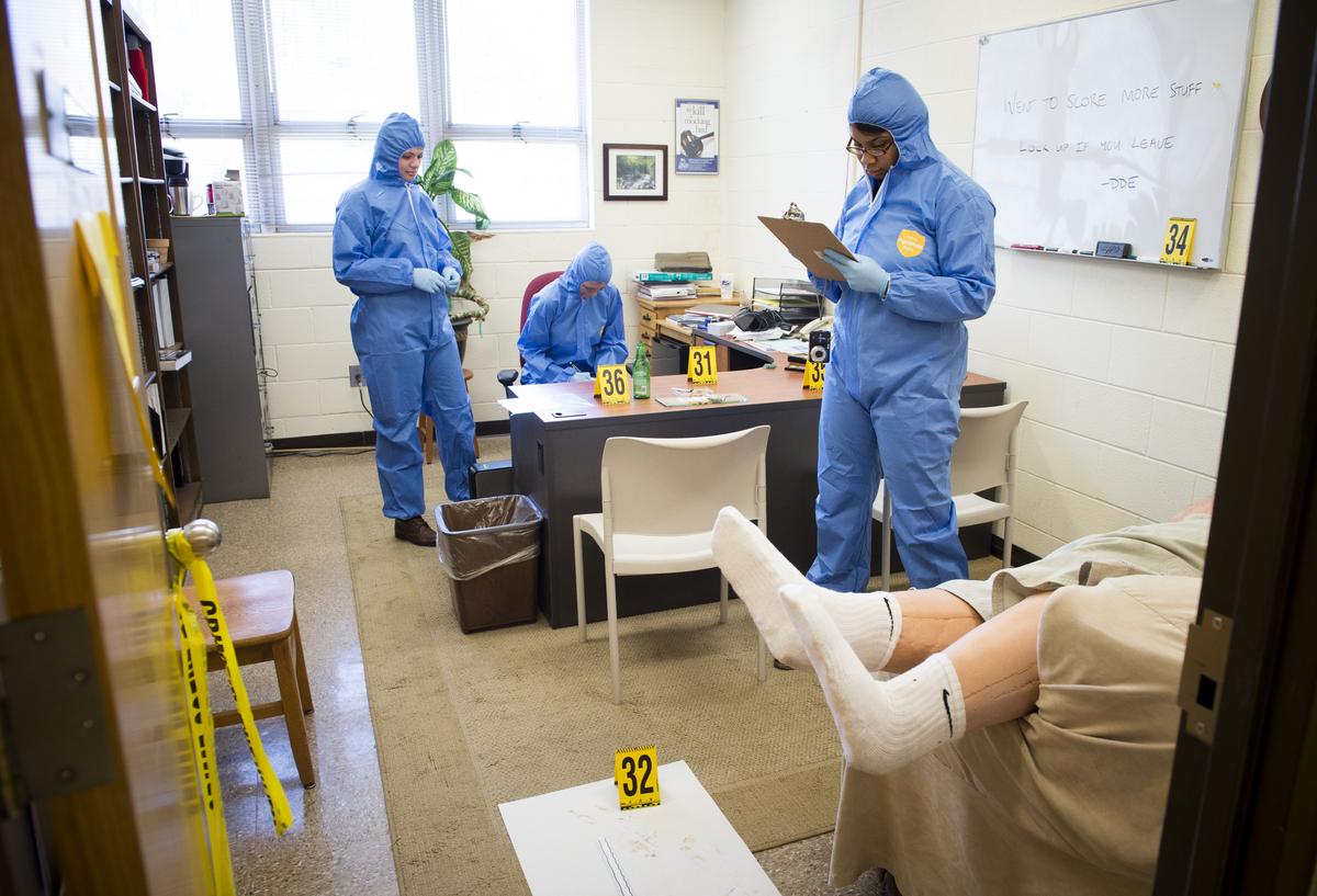 Students in hazmat suits looking over a fake crime scene taking notes