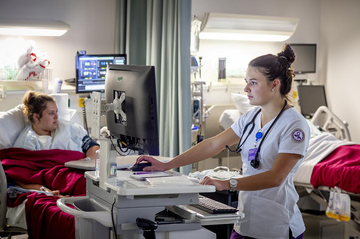 Student nurse practitioner working on a computer in a hospital room with patients in the background laying in beds