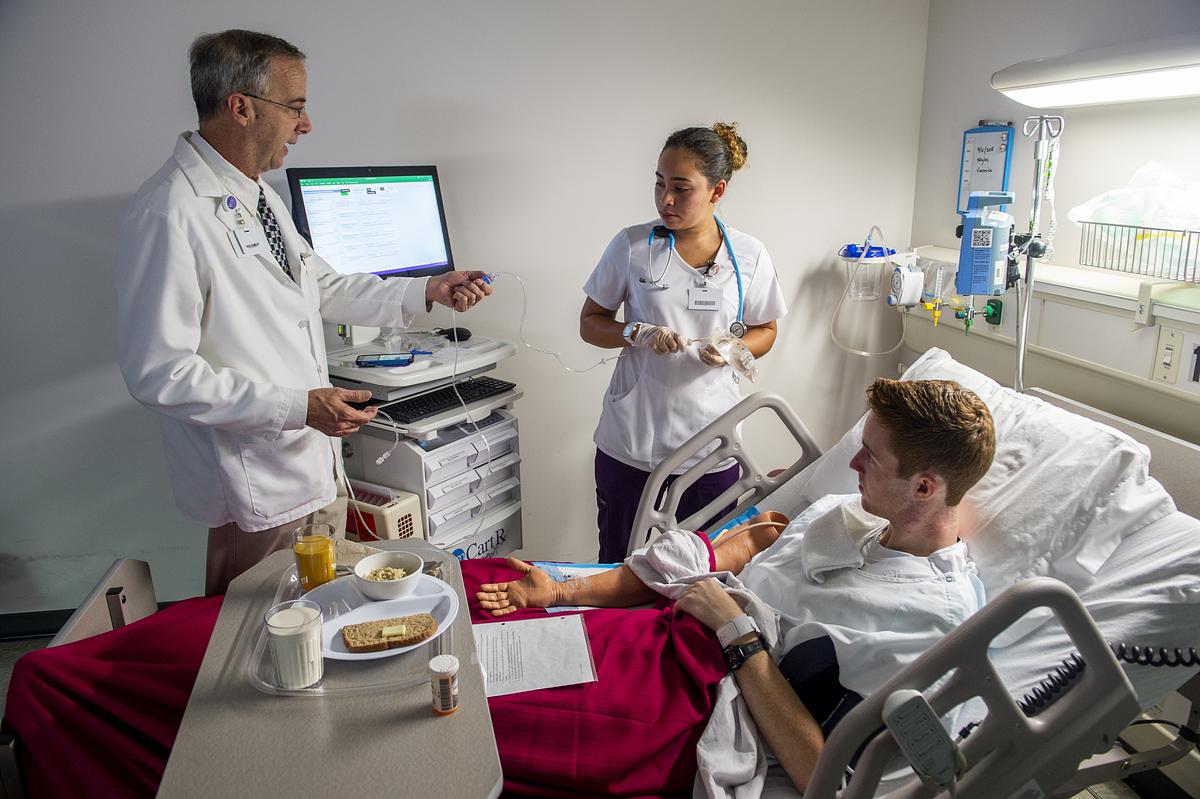 Doctor in white coat showing tablet to nurse while patient eats in hospital room.