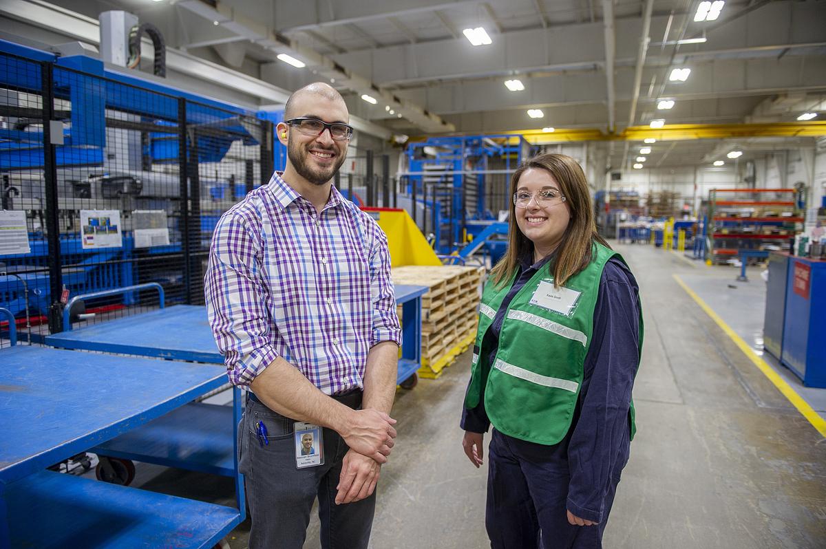 Man and younger woman standing in a warehouse wearing protective glasses.
