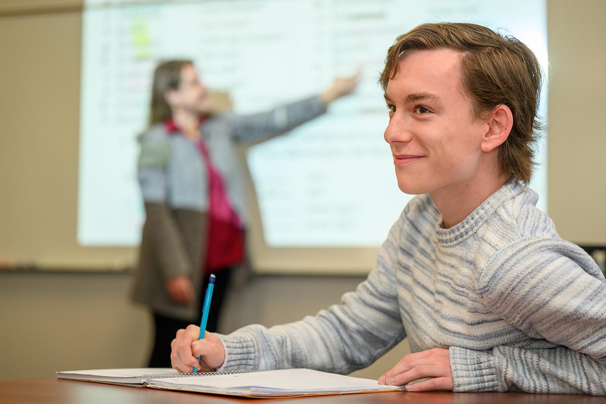 Student writing at a desk smiling while a teacher instructs the class on a whiteboard in the background