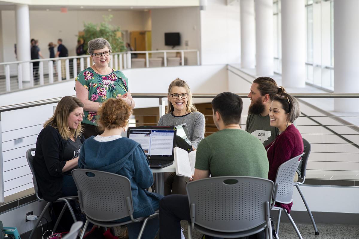 Professor standing and talking to a group of students sitting around a table working on their computers