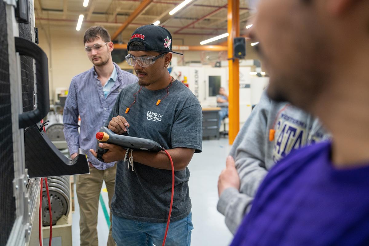 Technician wearing cap and glasses demonstrates electrical equipment to three colleagues in industrial workshop setting.