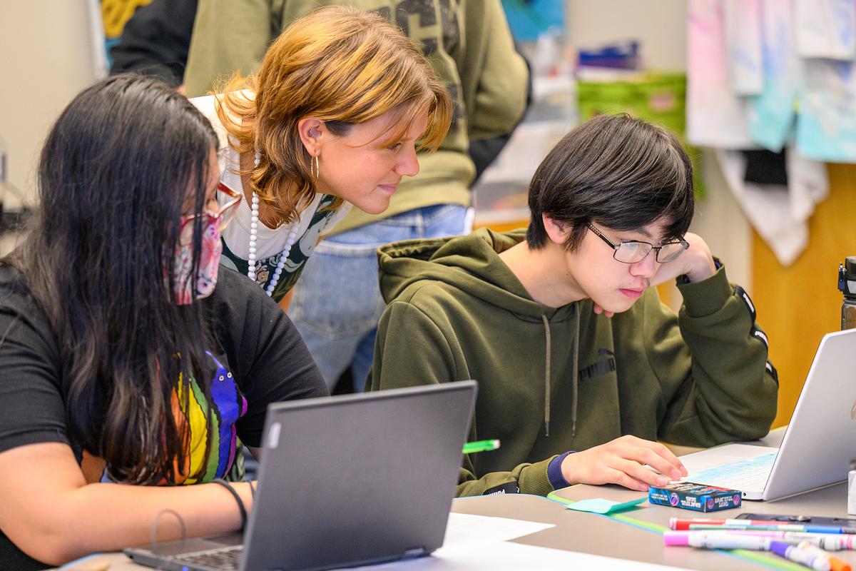 Teacher leaning over a desk with two students as they work on their laptops