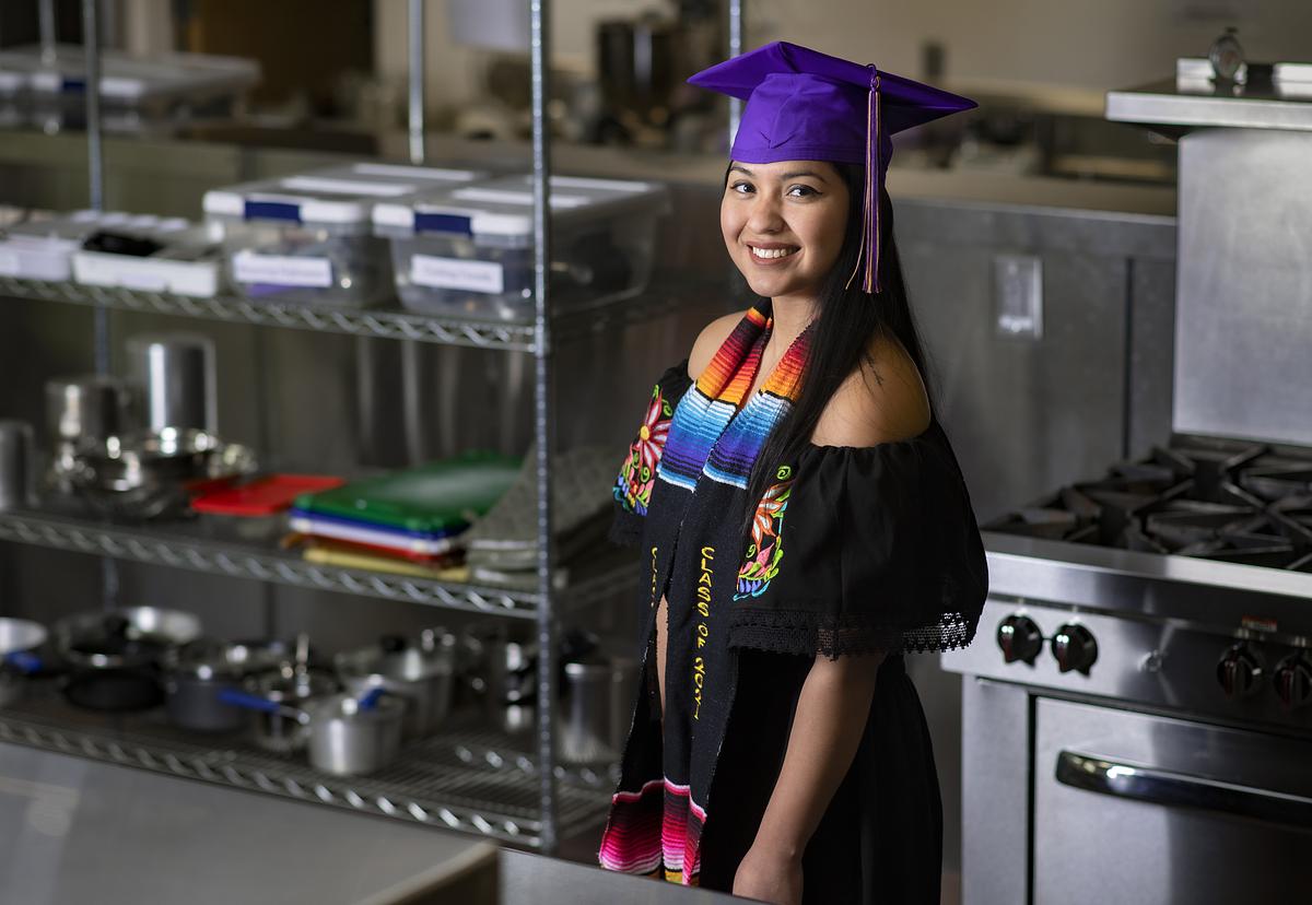 Graduate student Tierra Martinez smiles for the camera in a kitchen with her graduation cap and gown
