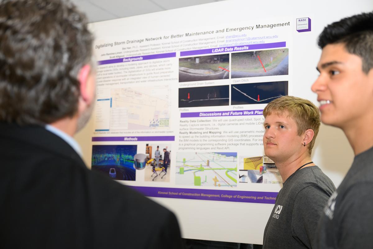 Students listen to a teacher as they go over a plan on a poster