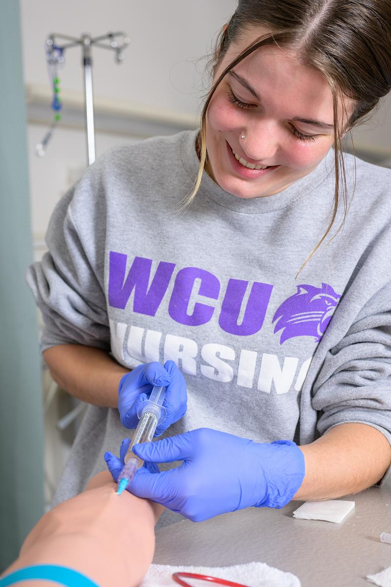 Nursing student in grey sweatshirt practices giving needle on test dummy