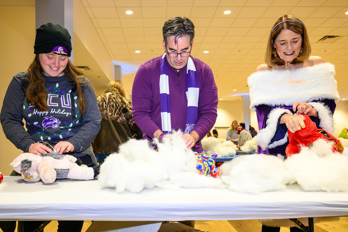 Three people stuffing teddy animals with white stuffing at a table during an indoor holiday event.