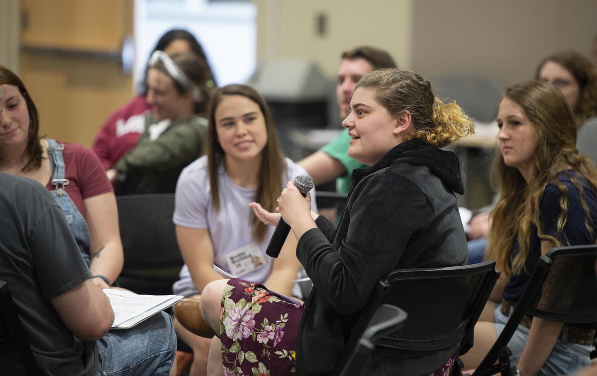 Student holding a microphone and speaking in a chair with other students sitting and listening to her