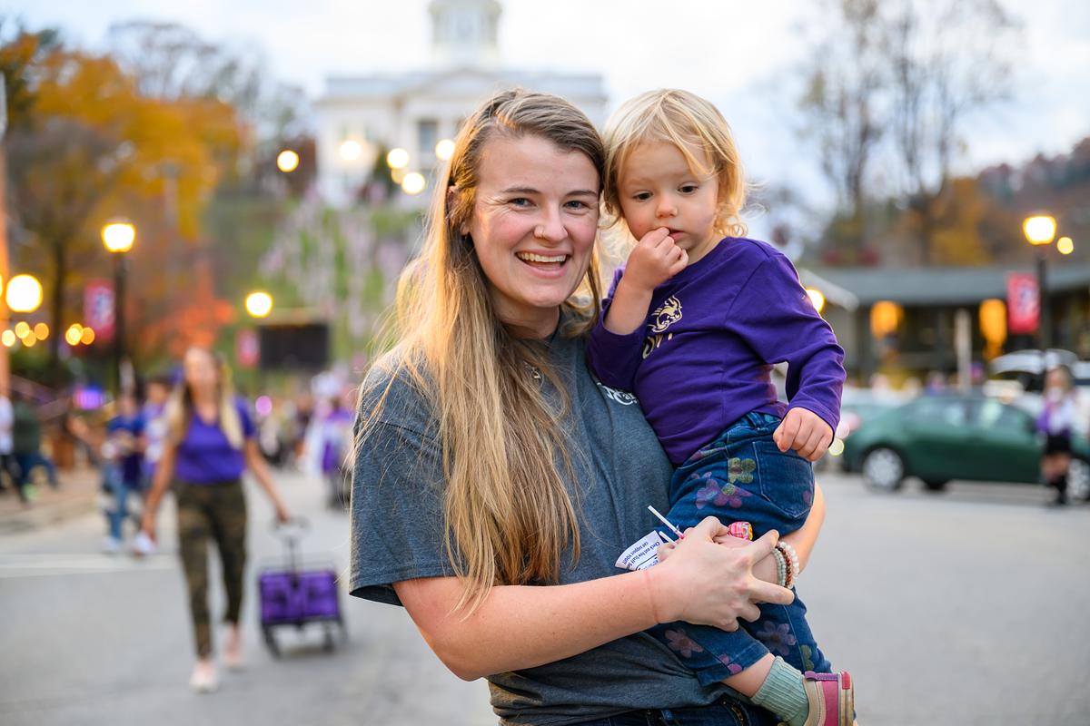 Parent holding a child and smiling for the camera in the homecoming parade alumni party