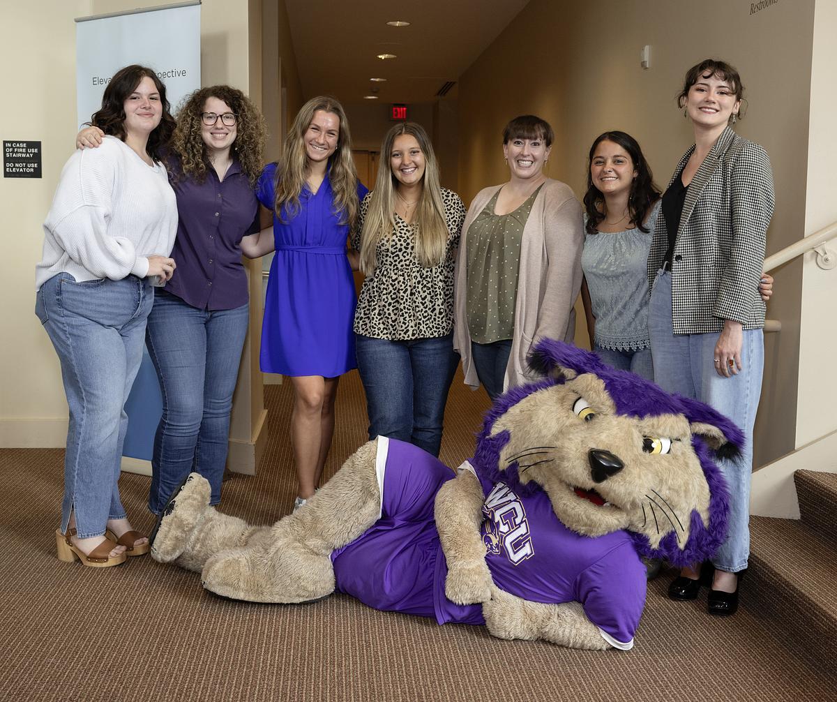 Seven women pose with a large purple and tan catamount mascot in an indoor hallway.
