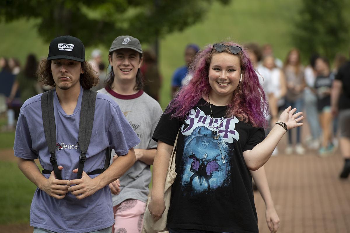 Girl with light red hair waves and smiles at the camera while walking with two friends