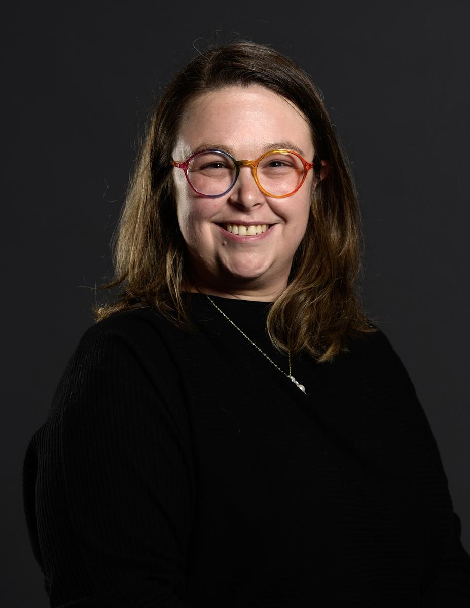 Chequita Brooks, a woman wearing multicolor-framed glasses and black shirt smiling at camera against dark background