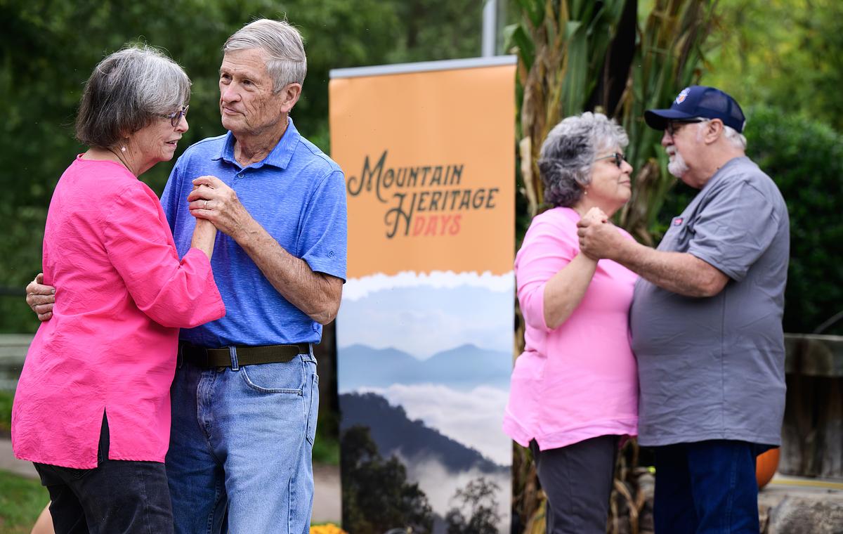 Four older adults in casual clothing conversing outdoors near a Mountain Heritage sign and green foliage.