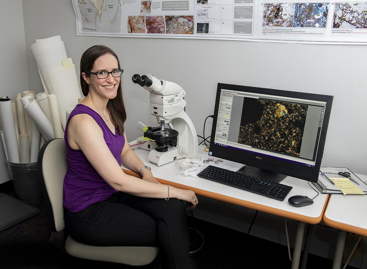Amy Fagan smiling for the camera as she studies some material under a microscope that is hooked up to a computer monitor