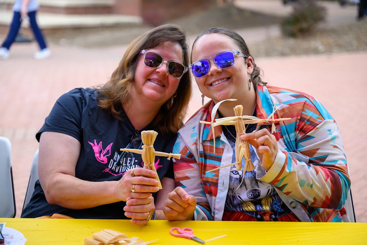 Two women wearing sunglasses holding wooden corn cob holders at outdoor event with yellow table.