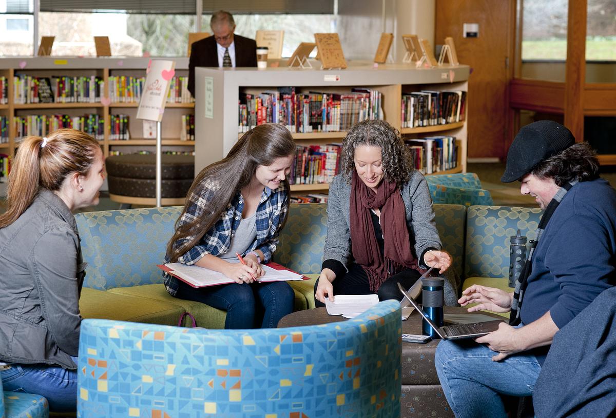 Students and teacher sitting around a lounge smiling and talking as they work together over documents 