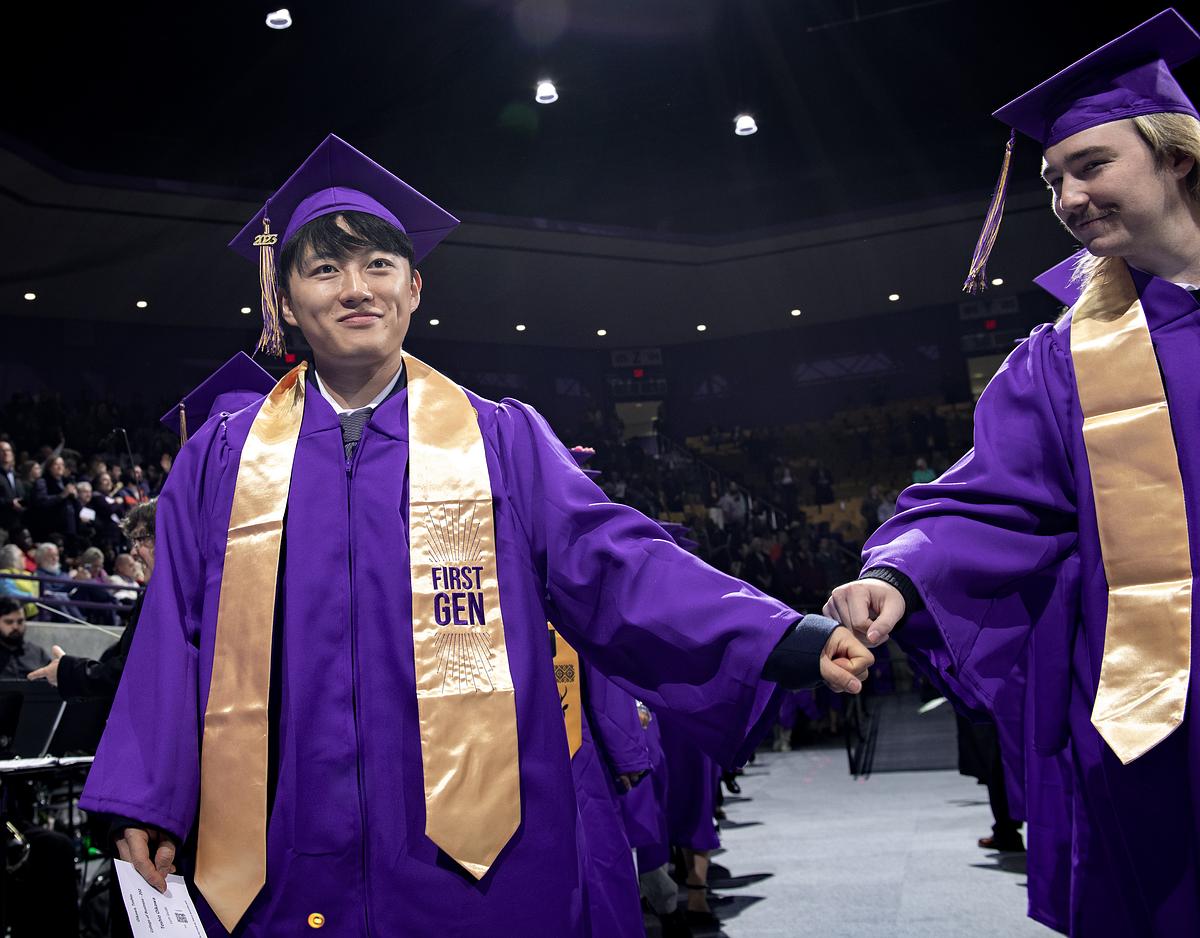 Two students in purple gowns and gold First Gen stoles fist bump at graduation