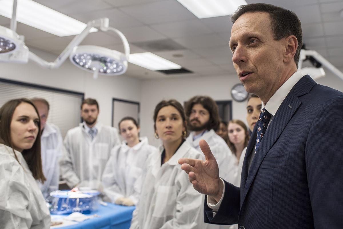 Picture of man in suit talking in a lab surrounded by students in white lab coats