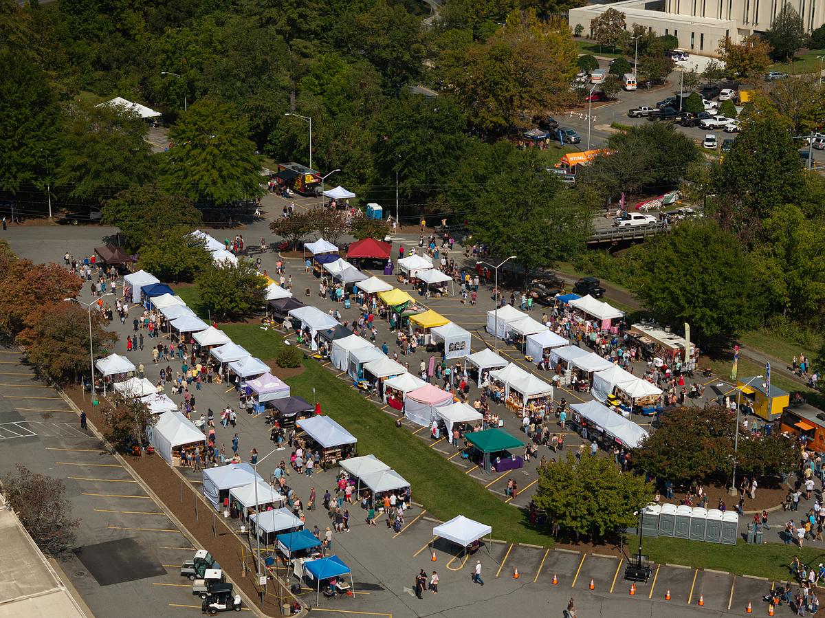 Aerial view of outdoor farmer's market with white vendor tents and crowds shopping in tree-lined venue