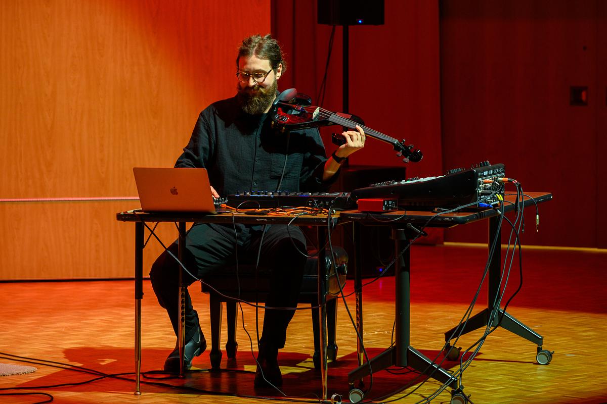 Musician sits at a desk with laptop and multiple musical equipment and instruments at a recital