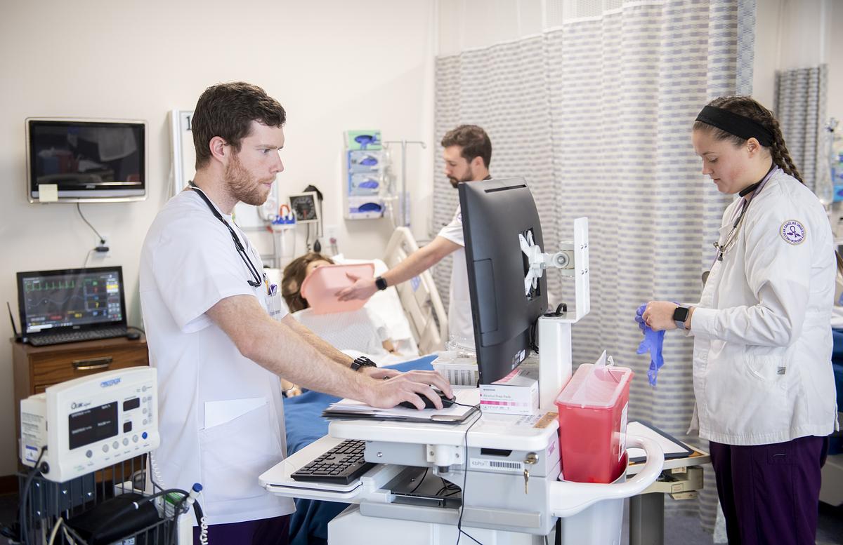 Medical professionals in clinical setting working at computer workstation with patient examination equipment nearby.
