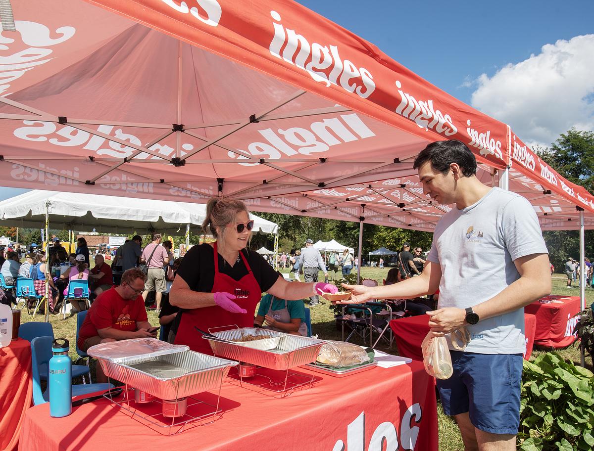 Vendor serving food at outdoor market tent while customer receives meal under red Moms Meals branded canopy