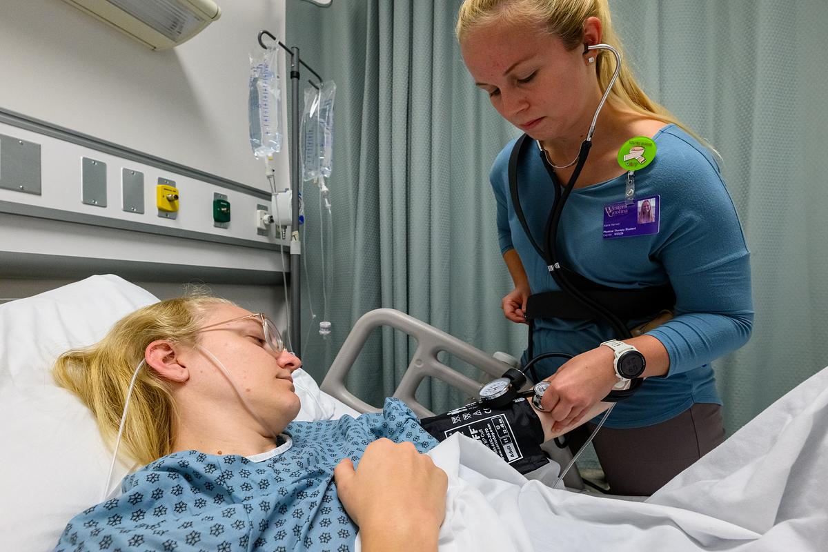 Student in a hospital gown and bed gets their blood pressure taken by a nurse