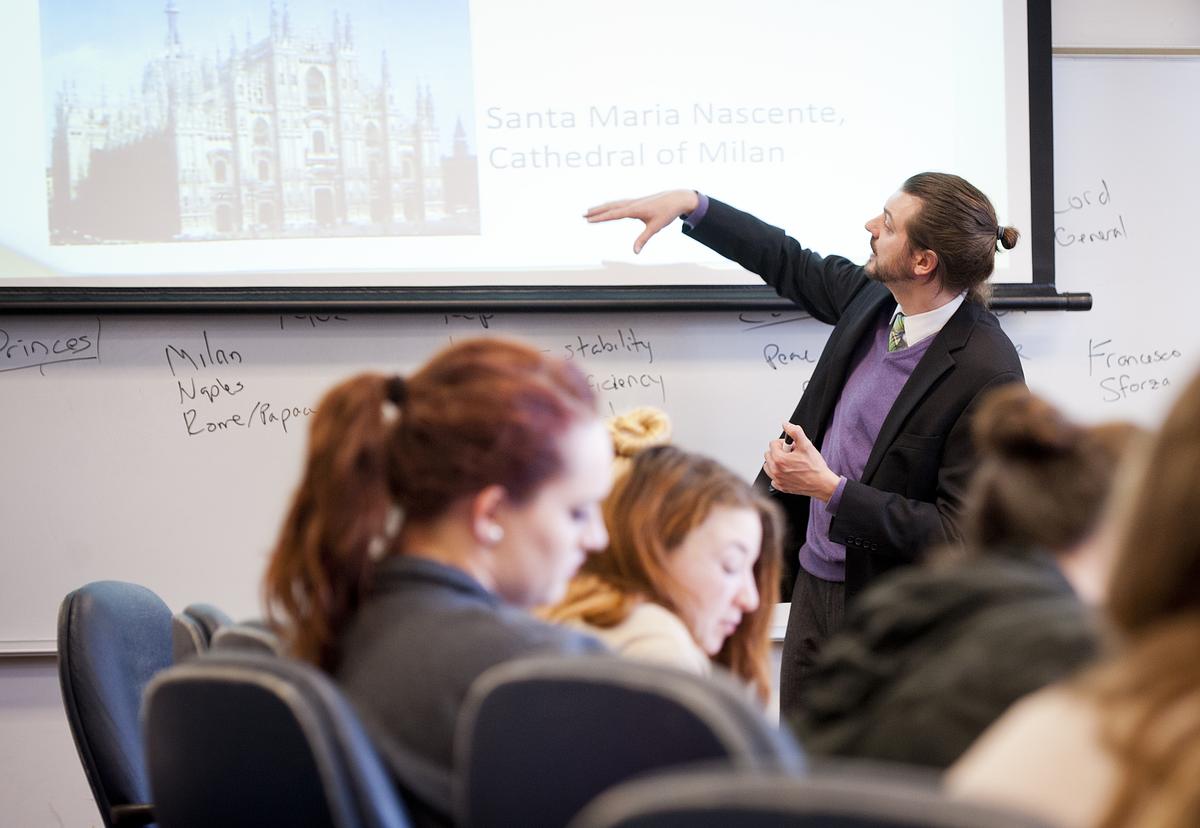 Professor sitting at a long desk with multiple students talking and working on their computers