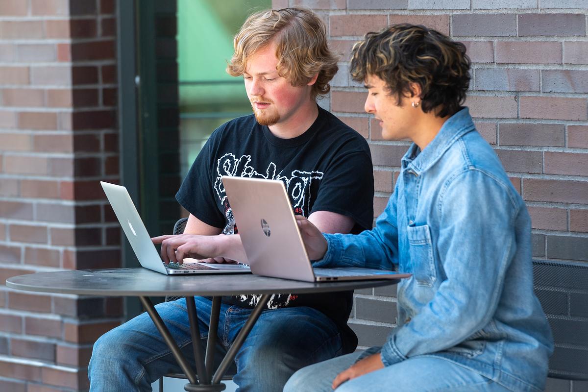 Two students sit at a table smiling and working on their laptops outside