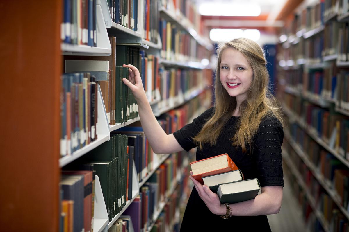 English graduate Anna Oates pulling books off of the shelf in the library and smiling for a photo