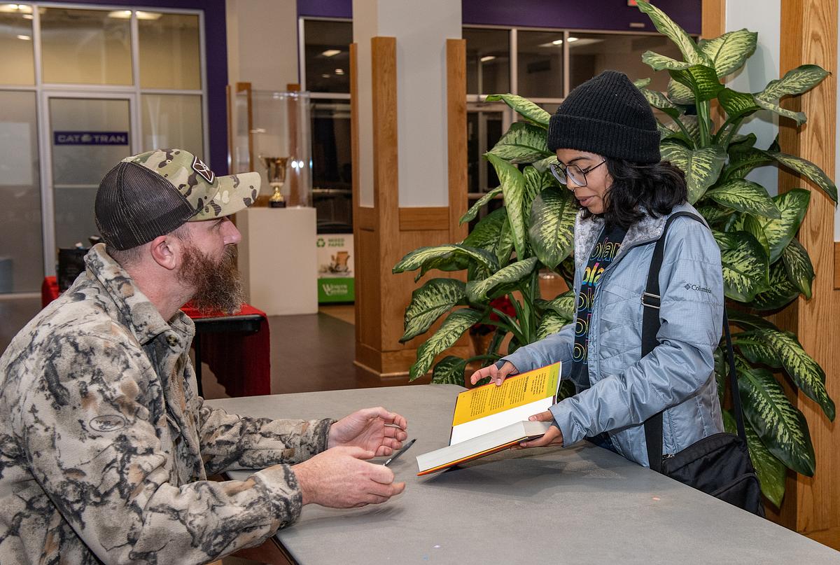 Student holding out a book to a army veteran at a table