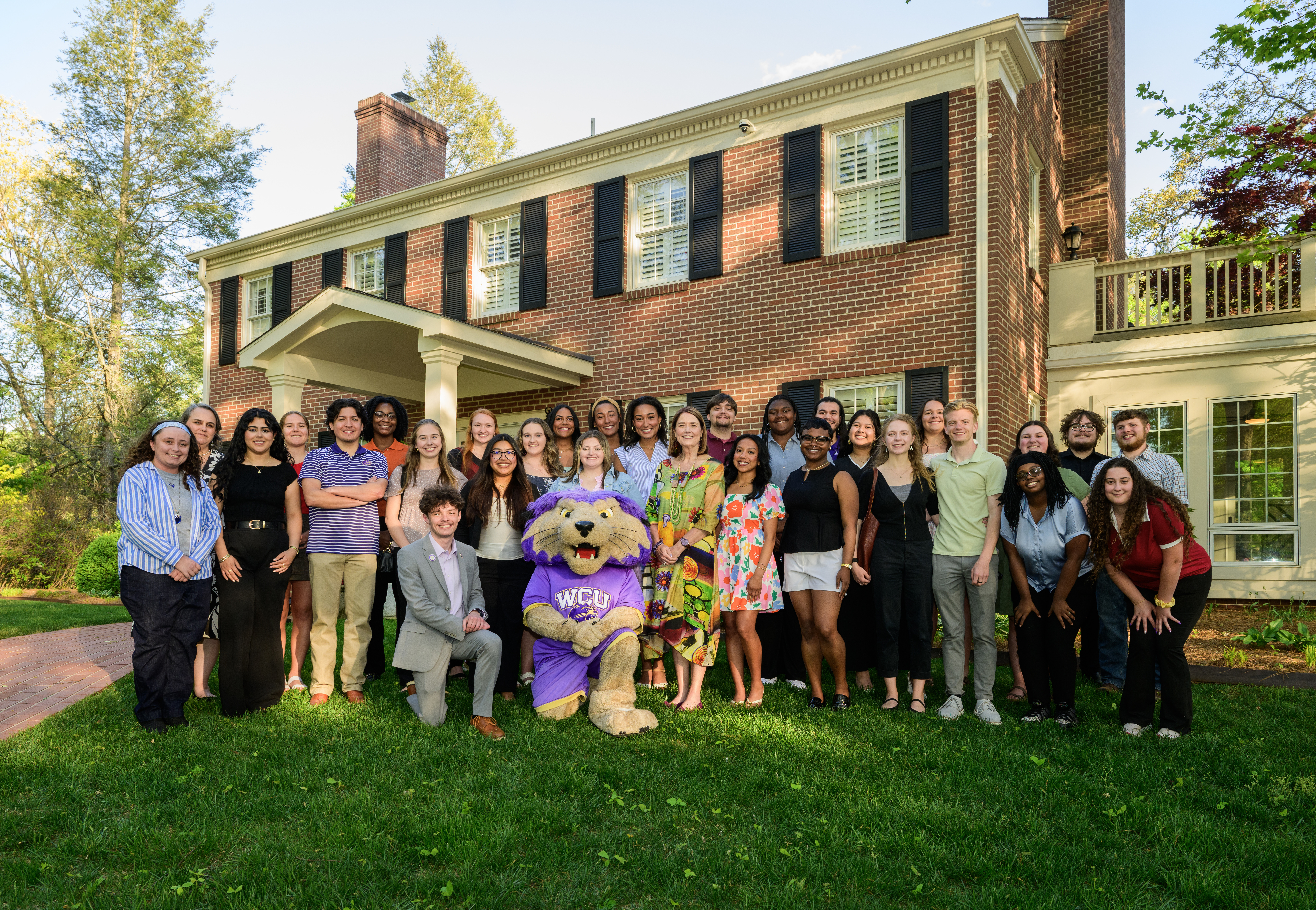 Forty-plus men and women pose with purple mascot costume on lawn before brick colonial house.
