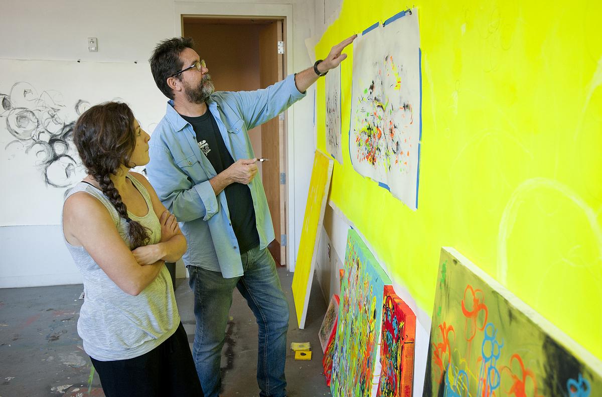 Teacher pointing out something on the painting in front of him and the student who is looking at it
