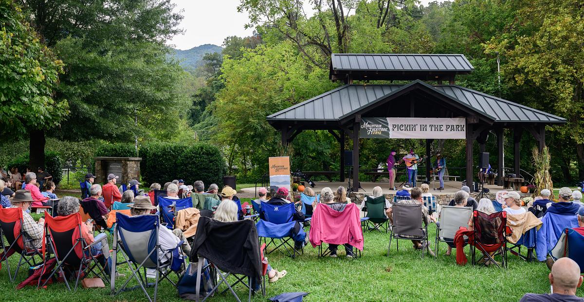 Outdoor picnic event in park with attendees seated in folding chairs under pavilion surrounded by trees.