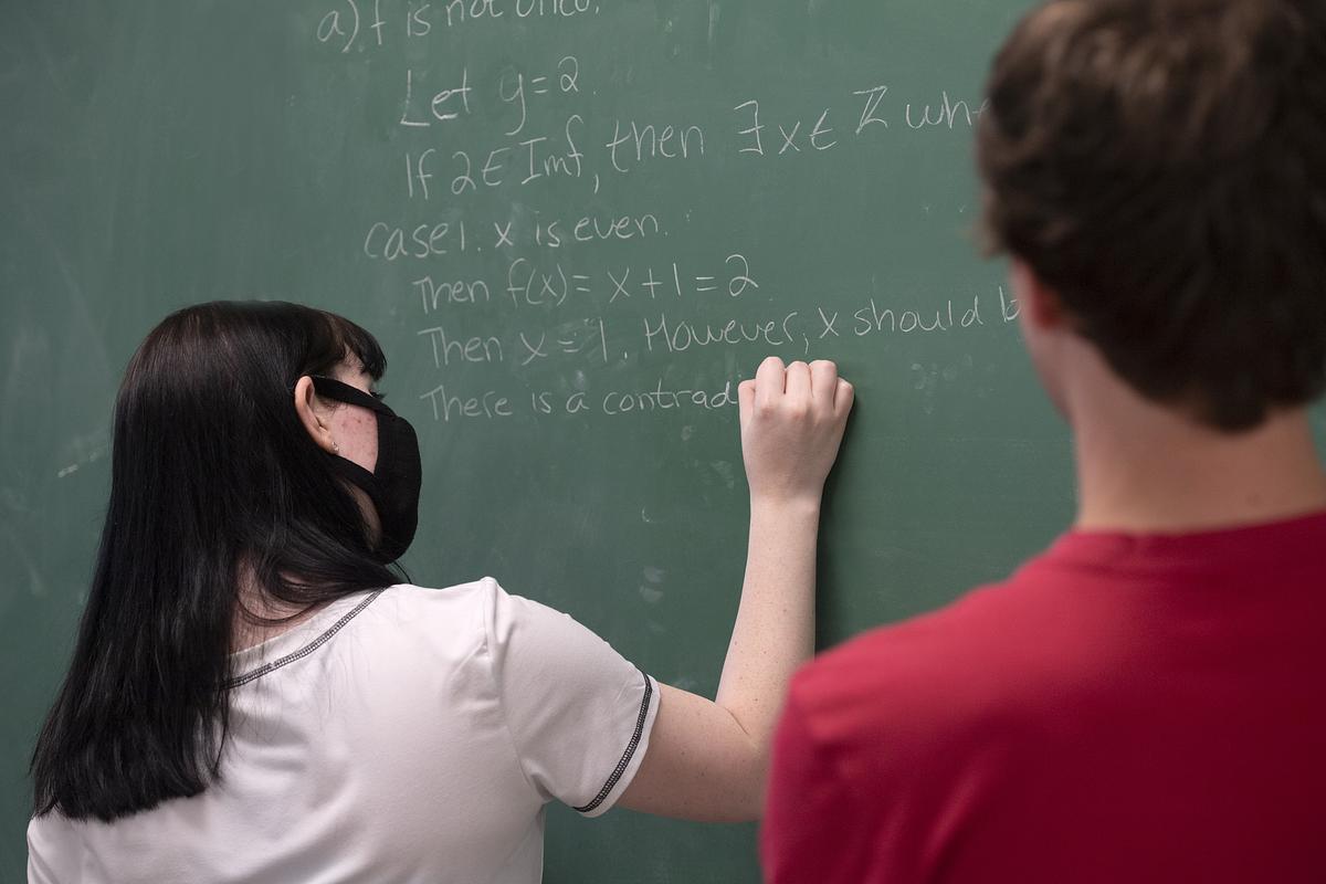 Student watches as another student solves a math problem on a chalkboard