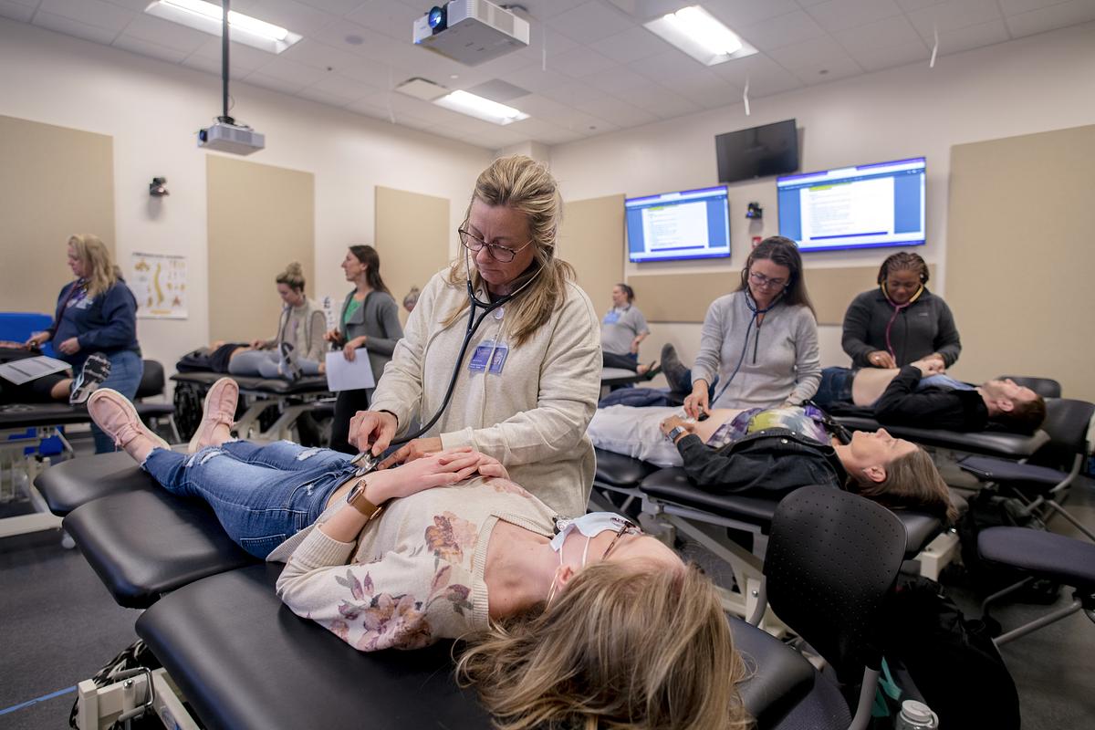 Medical instructor demonstrates abdominal palpation technique on student patient in classroom setting with other students observing.