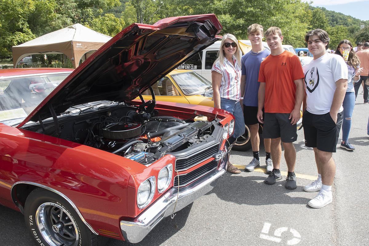 Group of five people posing beside red and yellow classic muscle cars with open hood at outdoor car show