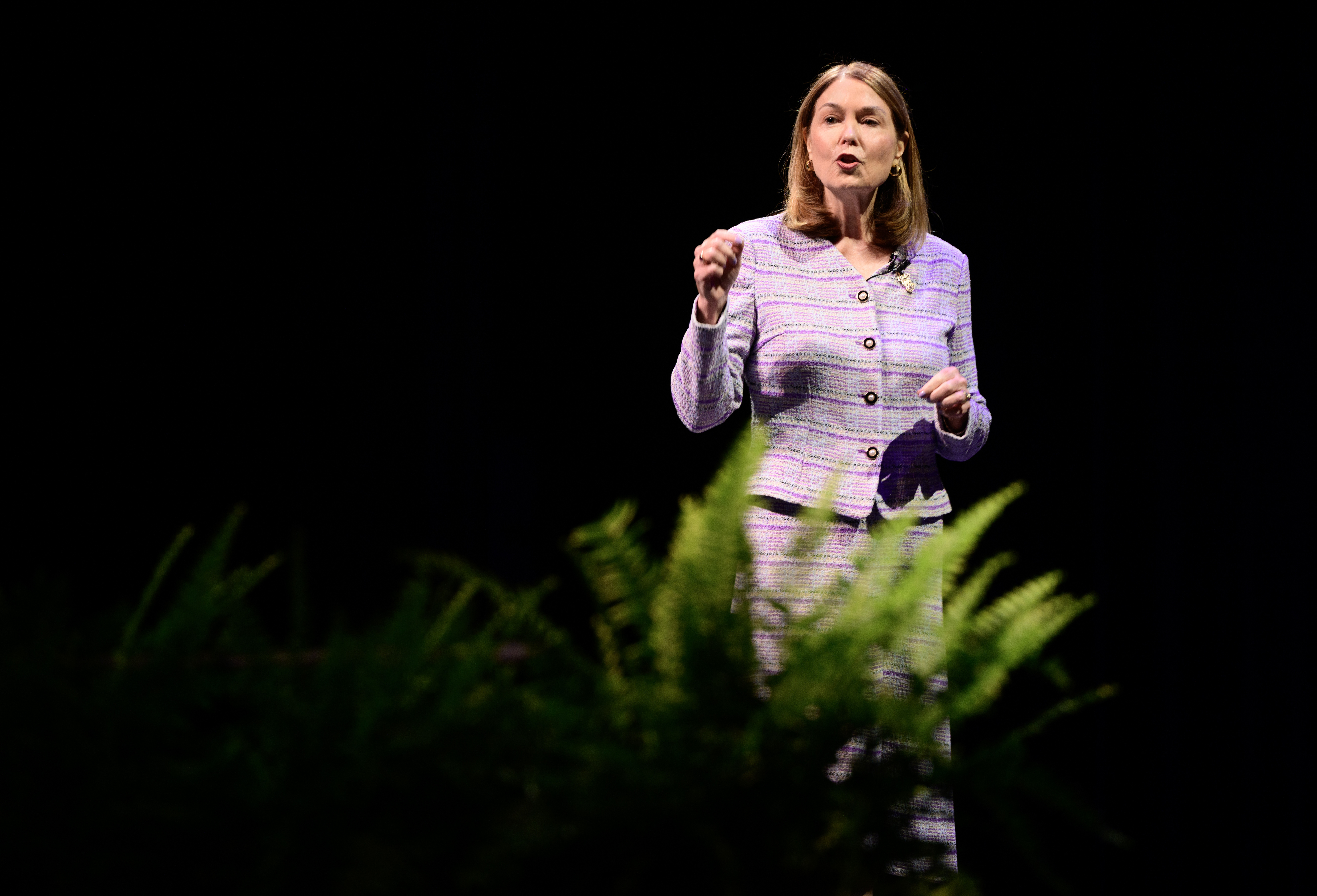 Woman in purple jacket speaking on stage with plants in foreground against dark background