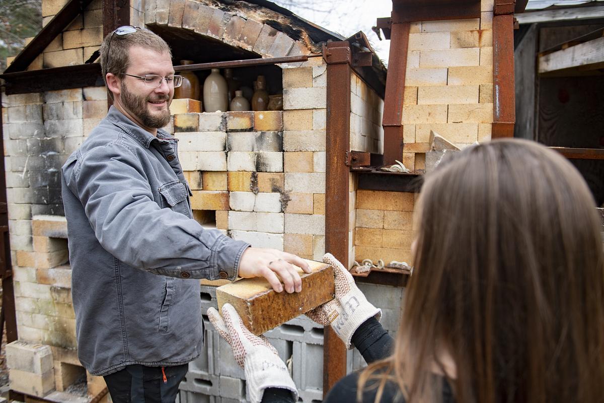 Faculty hands a student wearing gloves a brick from a recently fired kiln